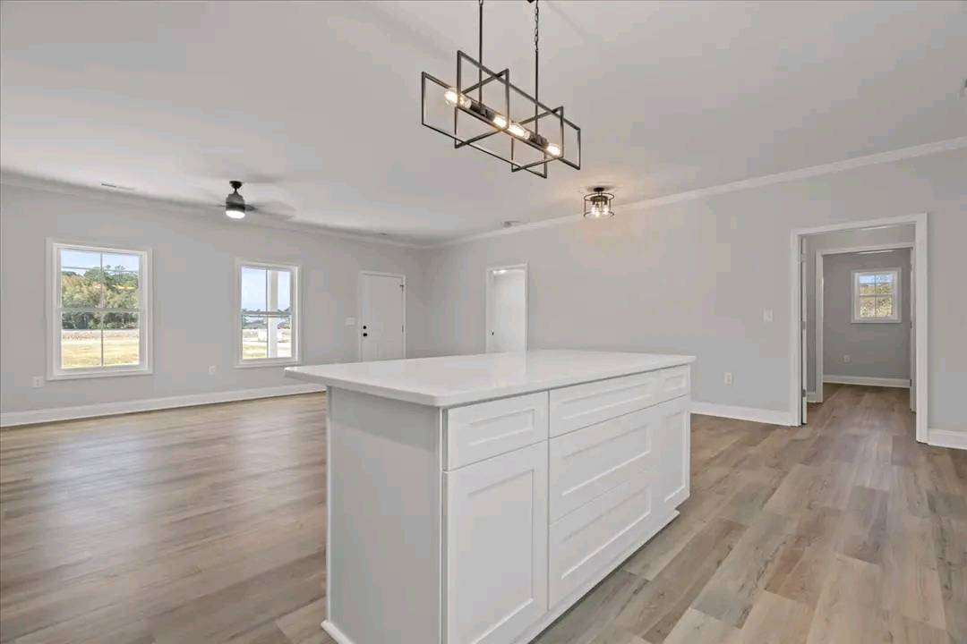 Spacious, modern open-plan room with light gray walls and wood flooring. A white kitchen island is centered under a geometric light fixture, near large windows.