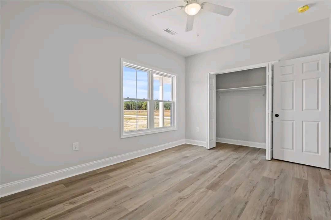 Bright, empty room with light wood flooring and soft gray walls. A large window shows a sunny view outside. An open closet and ceiling fan add to the spacious feel.