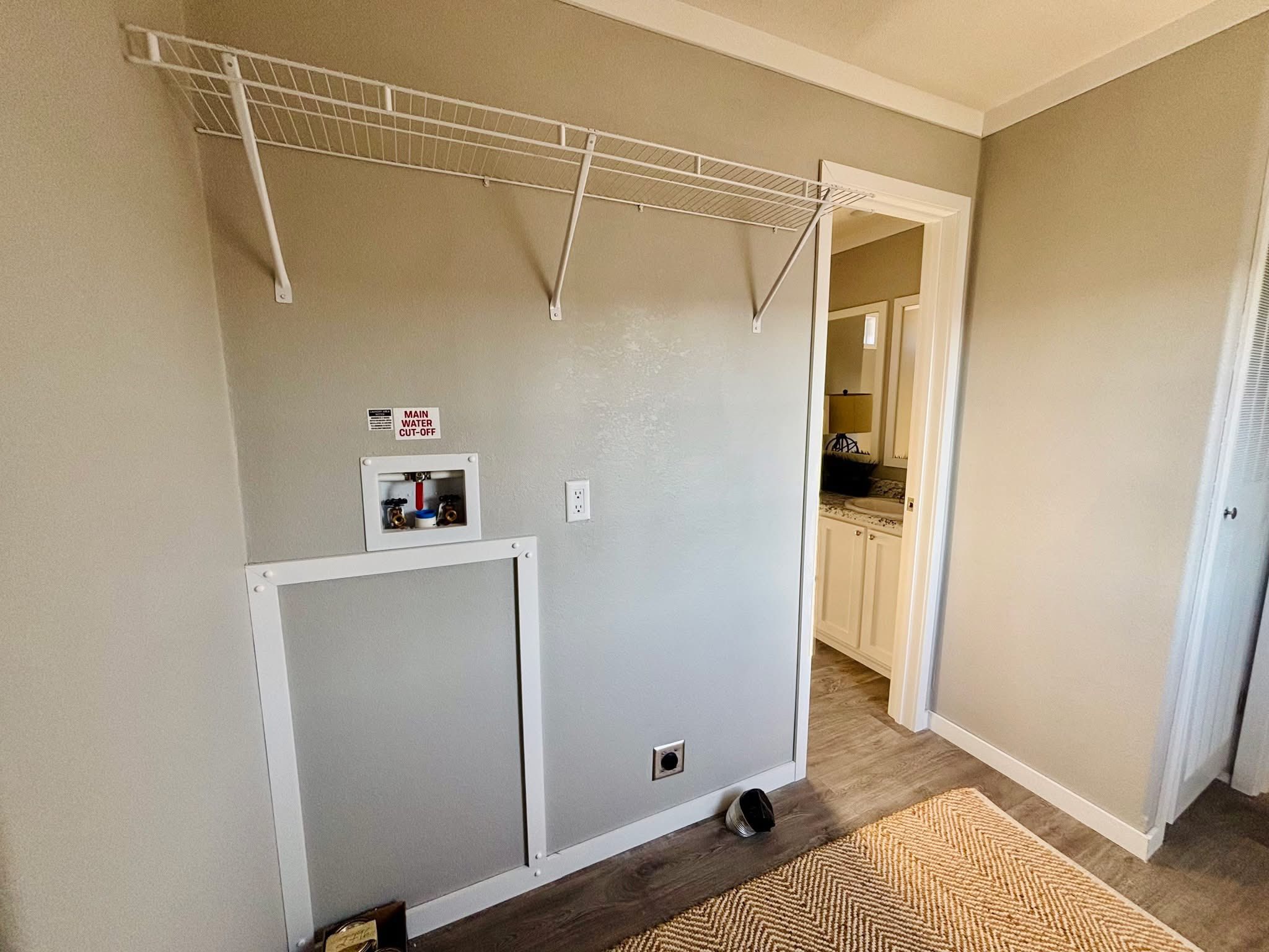 Room with light gray walls, featuring a white wire shelf above a plumbing setup with a "Main Water Shut Off" sign. Open doorway leads to a bathroom.