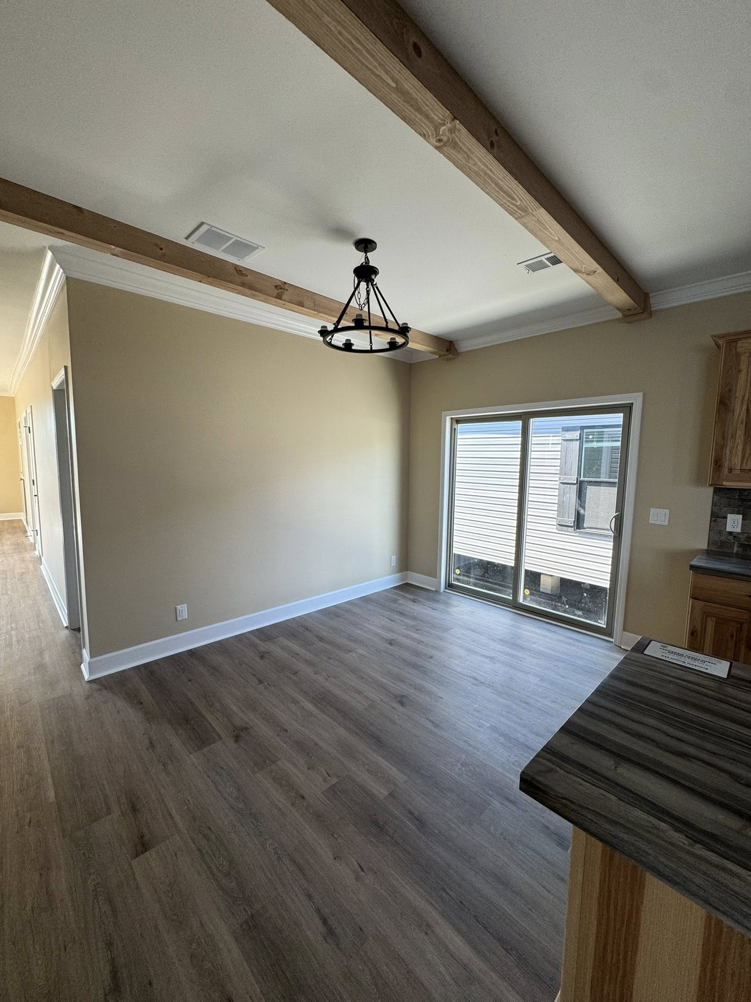 Spacious, empty dining area with light wooden floor, cream walls, exposed ceiling beams, and a black chandelier. Large sliding glass doors offer natural light.