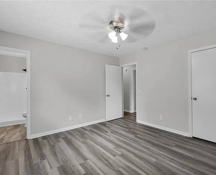 A spacious, empty room with light gray walls and wood laminate flooring. A ceiling fan with lights is in the center. Doors lead to a bright bathroom and hallway.