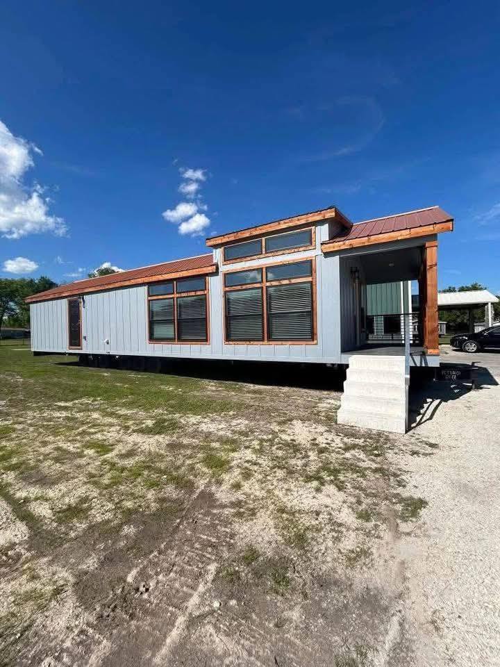 Modern tiny house with gray siding and large windows, set on a grassy plot under a clear blue sky. Concrete steps lead to a small porch.