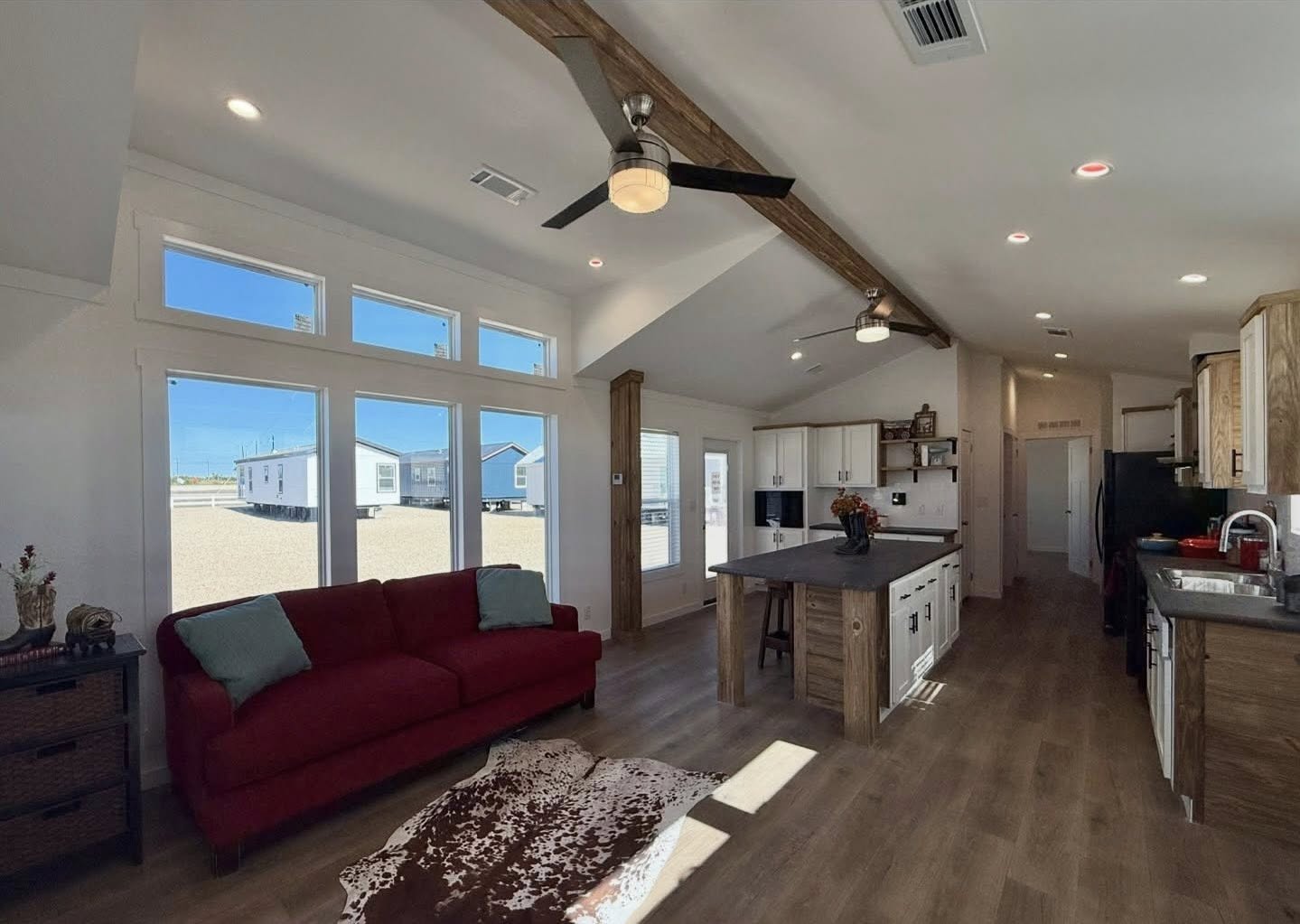 Spacious living room and kitchen with high ceilings, wooden beams, and a large window. Red sofa, cowhide rug, and modern kitchen island create a cozy yet contemporary feel.