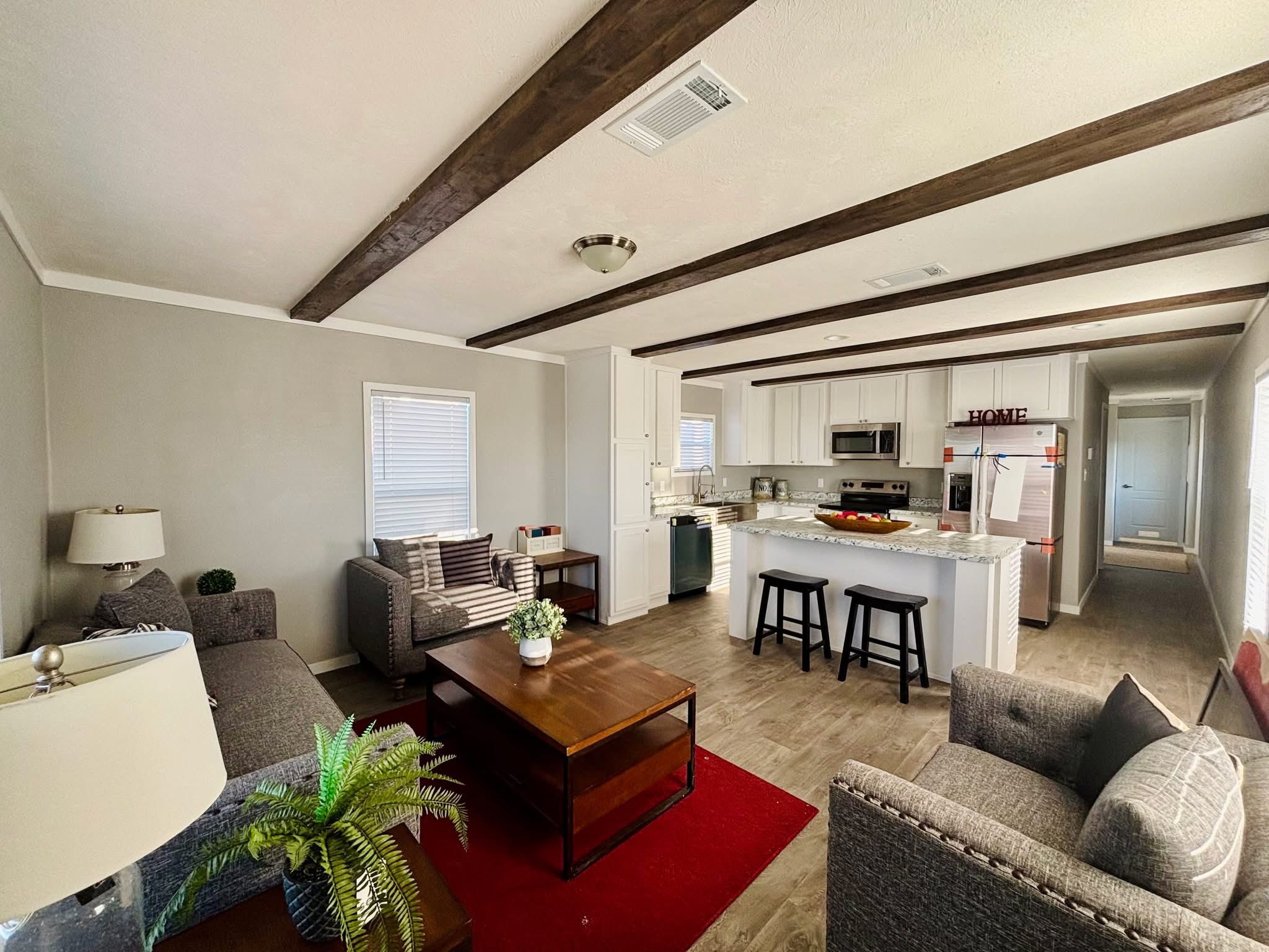 Modern open-plan living room and kitchen with gray sofas, a wooden coffee table on a red rug, and a kitchen island with black stools. Bright and cozy.