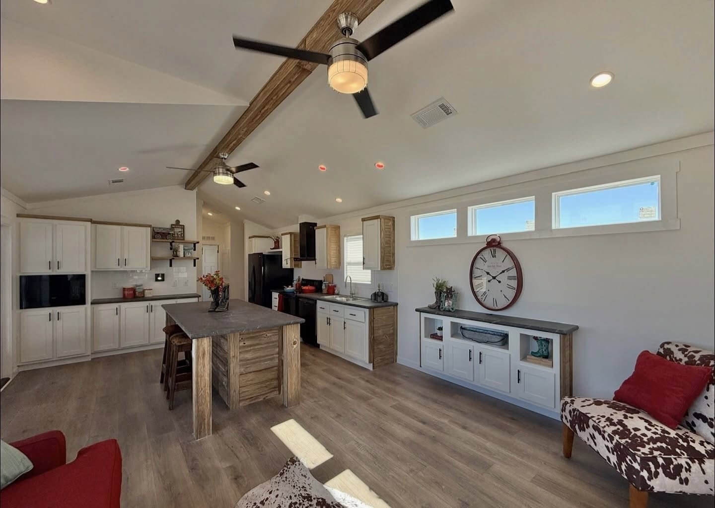 Spacious modern kitchen with rustic accents, featuring a wooden island, white cabinets, large wall clock, and ceiling fans. Bright and inviting atmosphere.