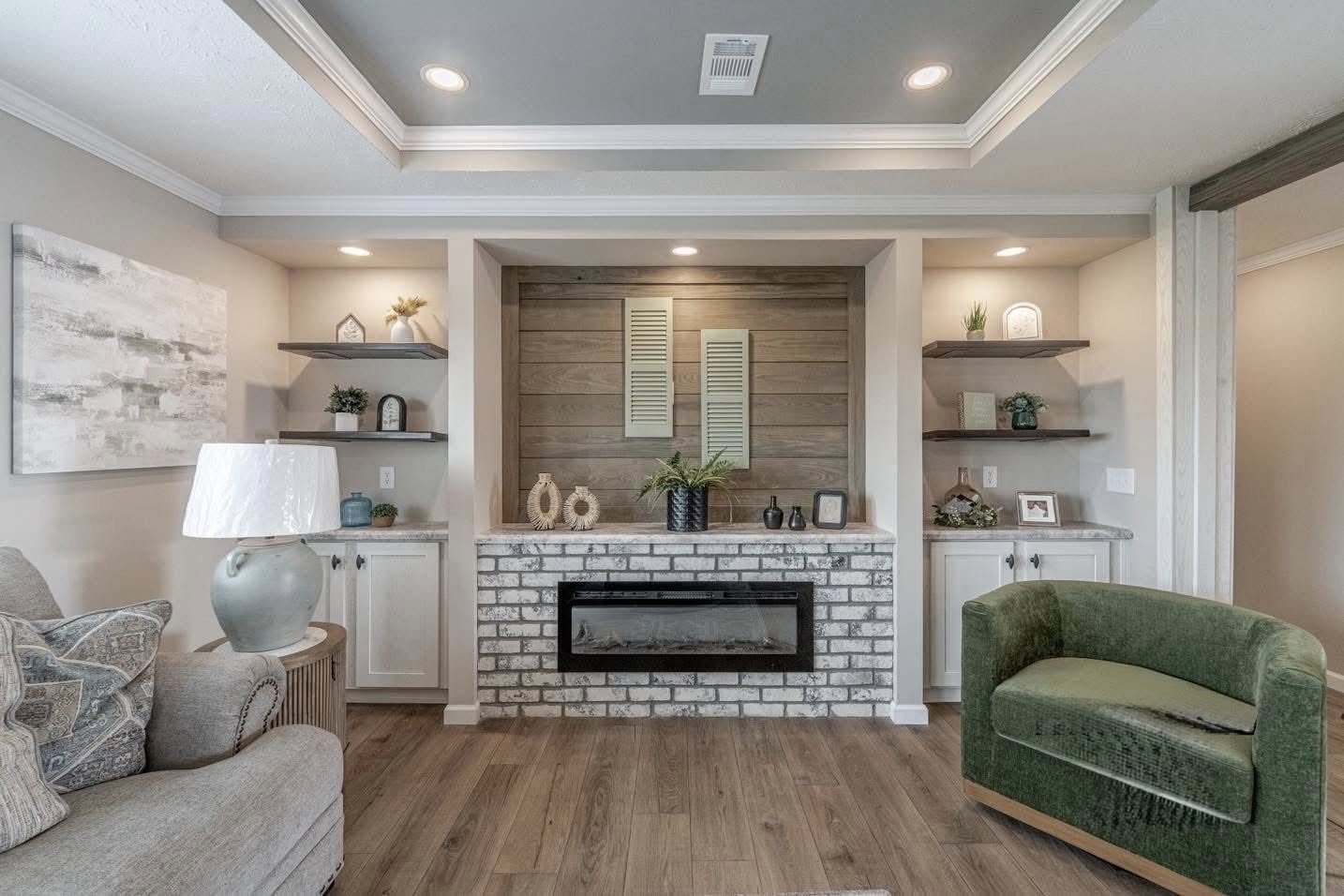Modern living room with a cozy vibe. Central brick fireplace, flanked by shelves with decor. Neutral tones, green armchair, and wood accents.
