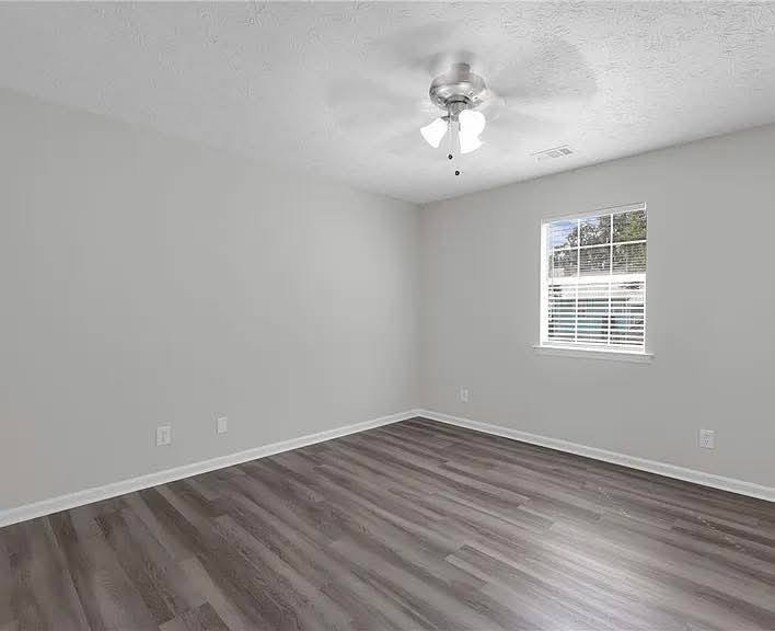 Empty room with gray walls and dark wooden flooring. A ceiling fan with lights hangs above, and a window on the right lets in natural light. Minimalist and neutral ambiance.