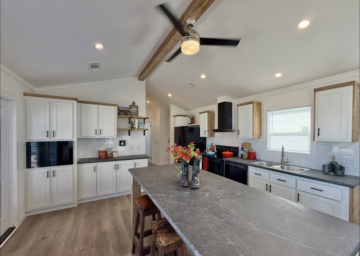 Spacious kitchen with a black marble island, bar stools, and flowers. White cabinets, wood accents, black appliances, ceiling fan, bright lighting.