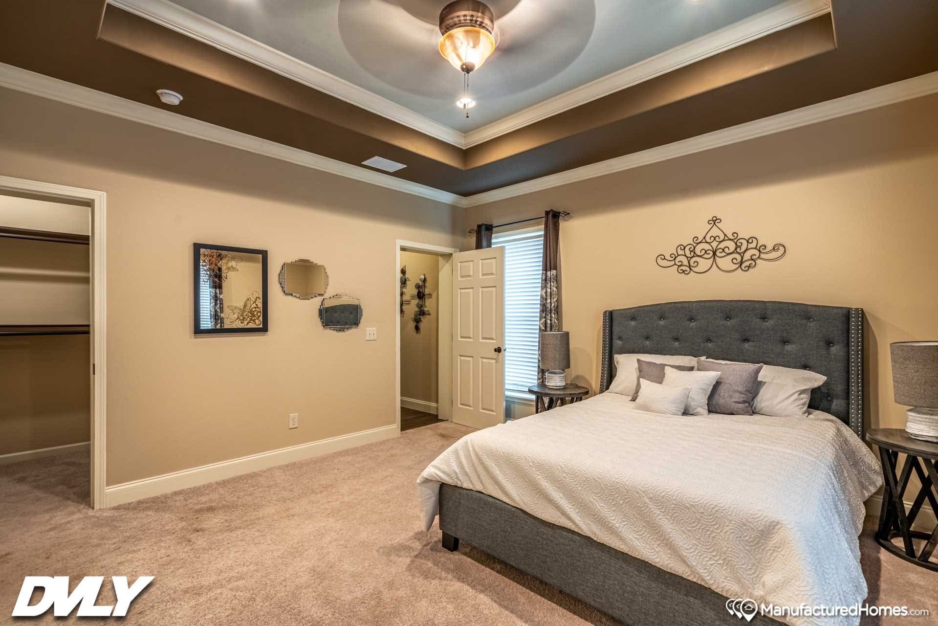 Elegant bedroom with a tray ceiling, ceiling fan, and soft lighting. Features a gray upholstered bed, two bedside tables with lamps, and decorative wall art.