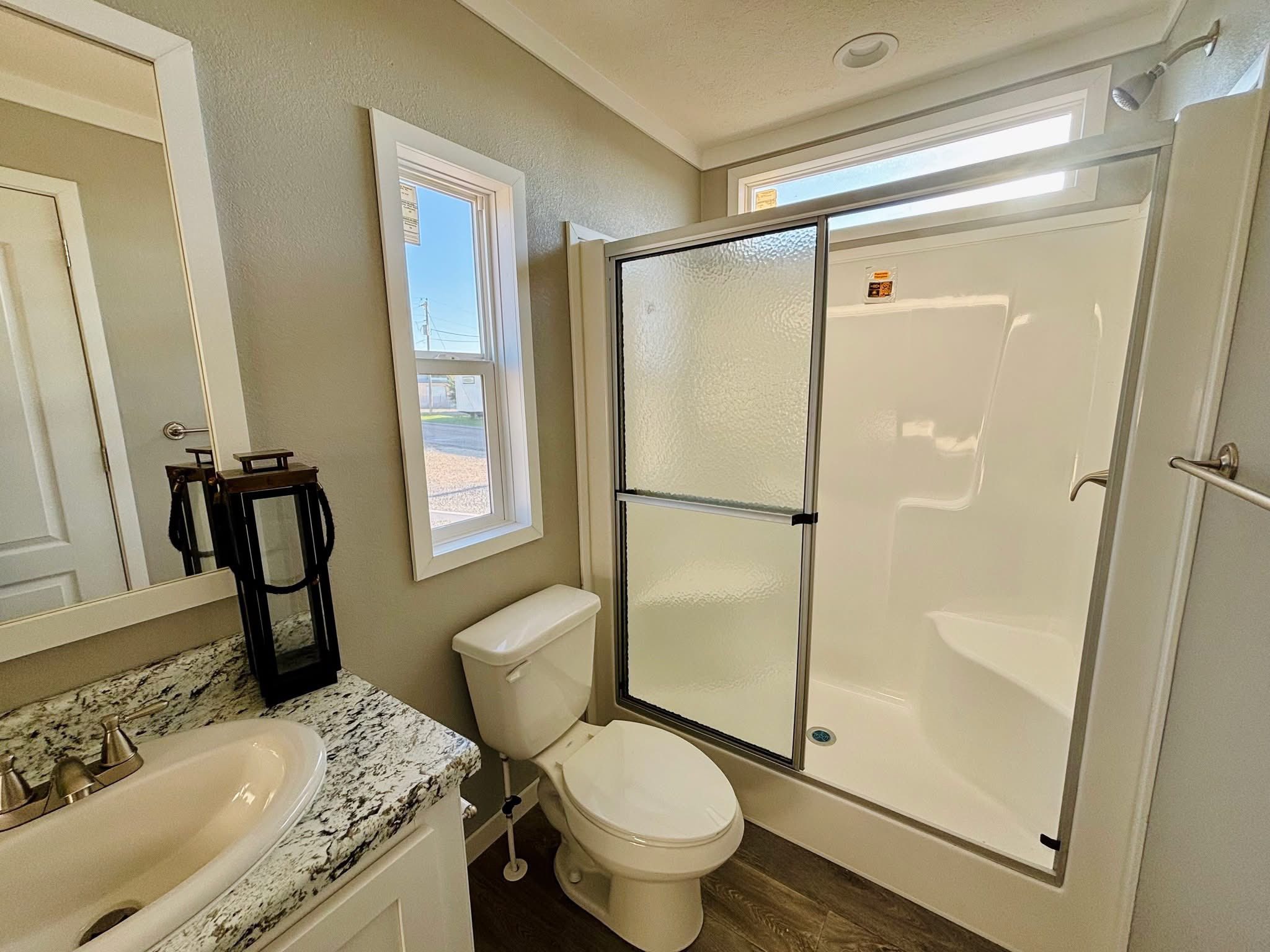 Compact modern bathroom with a granite countertop sink, white toilet, and a glass-enclosed shower. Natural light from two windows brightens the space.