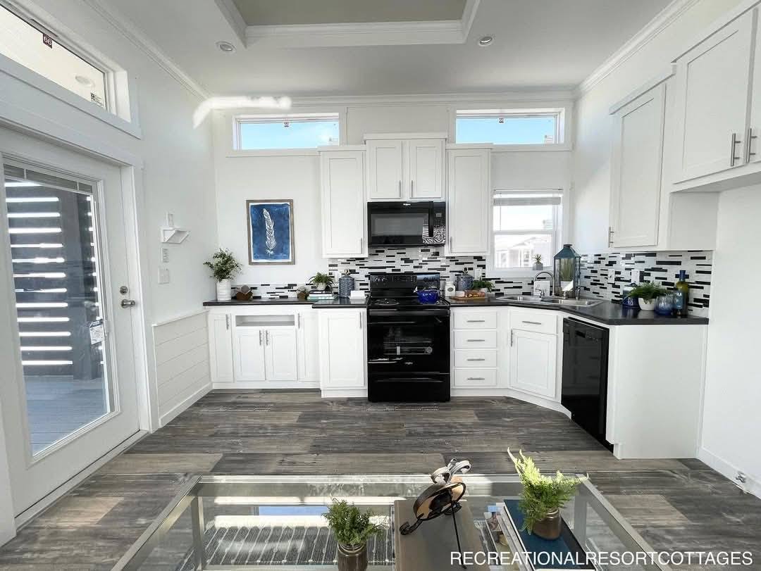 Modern kitchen with white cabinets, black appliances, and a mosaic tile backsplash. Sunlight streams in from high windows, creating an airy feel.