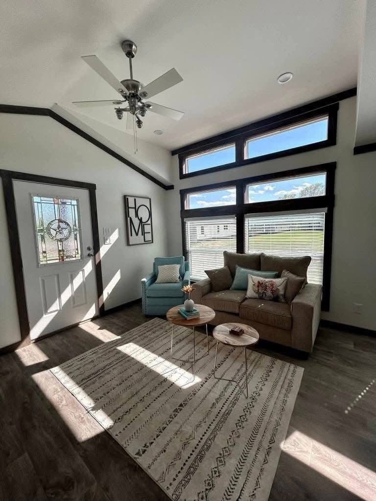 Modern living room with high ceilings, dark wood accents, and large windows. It features a beige sofa, blue chair, round tables, and a geometric rug.