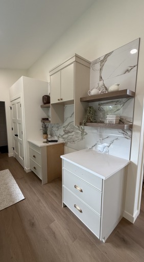 Modern kitchen with marble backsplash, white cabinets, wooden shelves, and decorative plants. Warm wood flooring adds a cozy, elegant touch.