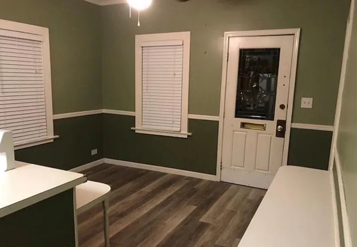 A small room with green walls, white trim, and wood flooring. Two windows with closed blinds and a white door are visible. The space looks clean and empty.
