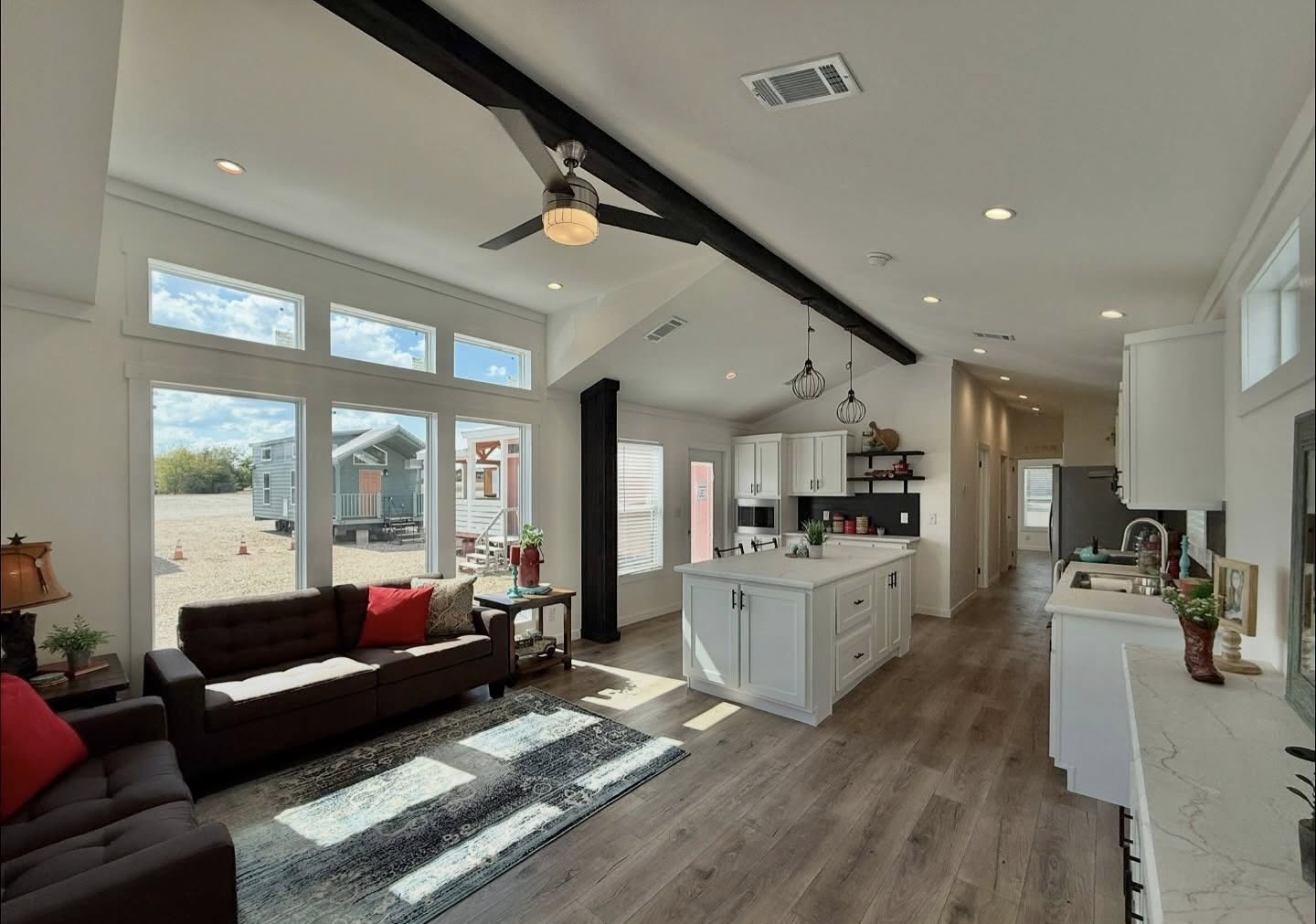Modern open-plan living area featuring large windows, a brown sofa with red cushions, and an island kitchen. Bright, airy, and inviting atmosphere.
