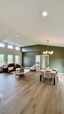 Spacious living room with high ceiling, modern chandelier, and large windows. Includes a dining area with a wooden floor and green accent wall, creating a cozy ambiance.