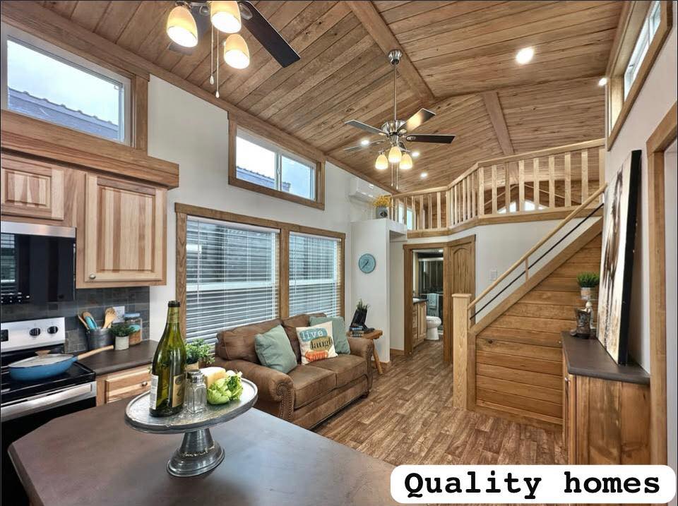 Cozy tiny home interior with wood paneling, a brown couch with colorful pillows, lofted ceiling, modern kitchen, stairs to loft, warm lighting.