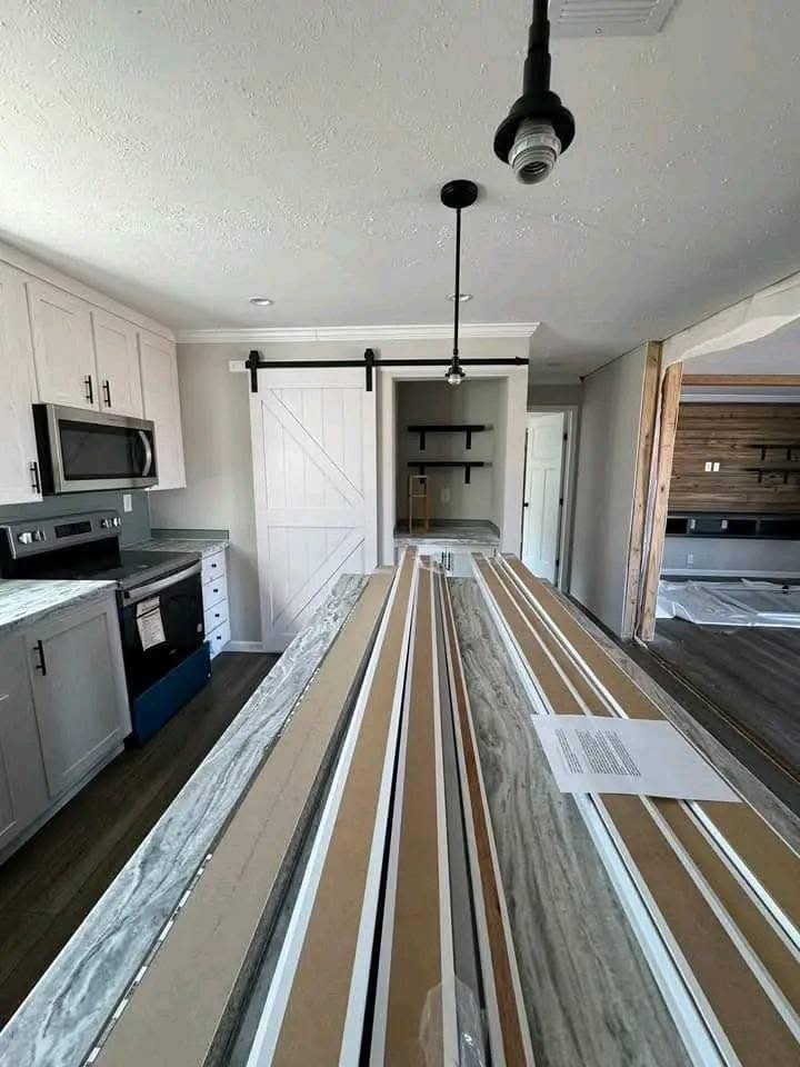 Modern kitchen interior with white cabinets, stainless steel appliances, and a large marble island. Pendant lights hang above, and a sliding barn door is visible in the background. The overall tone is sleek and contemporary.