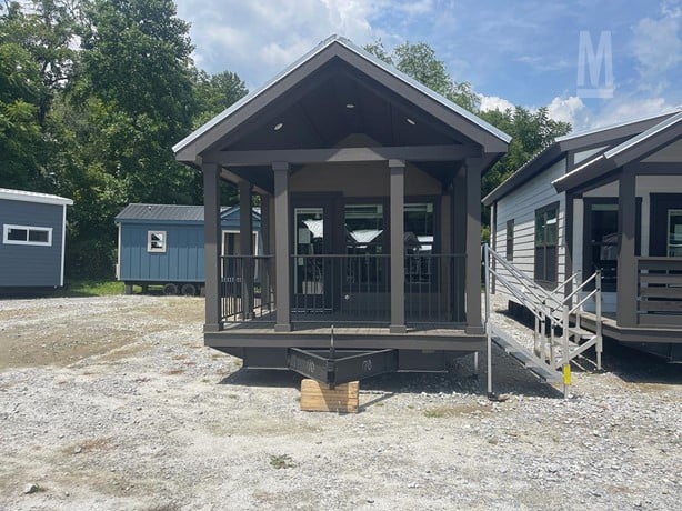 A small, modern tiny house with a dark wood exterior, set on a gravel lot. It features a covered porch and steps, surrounded by trees under a blue sky.
