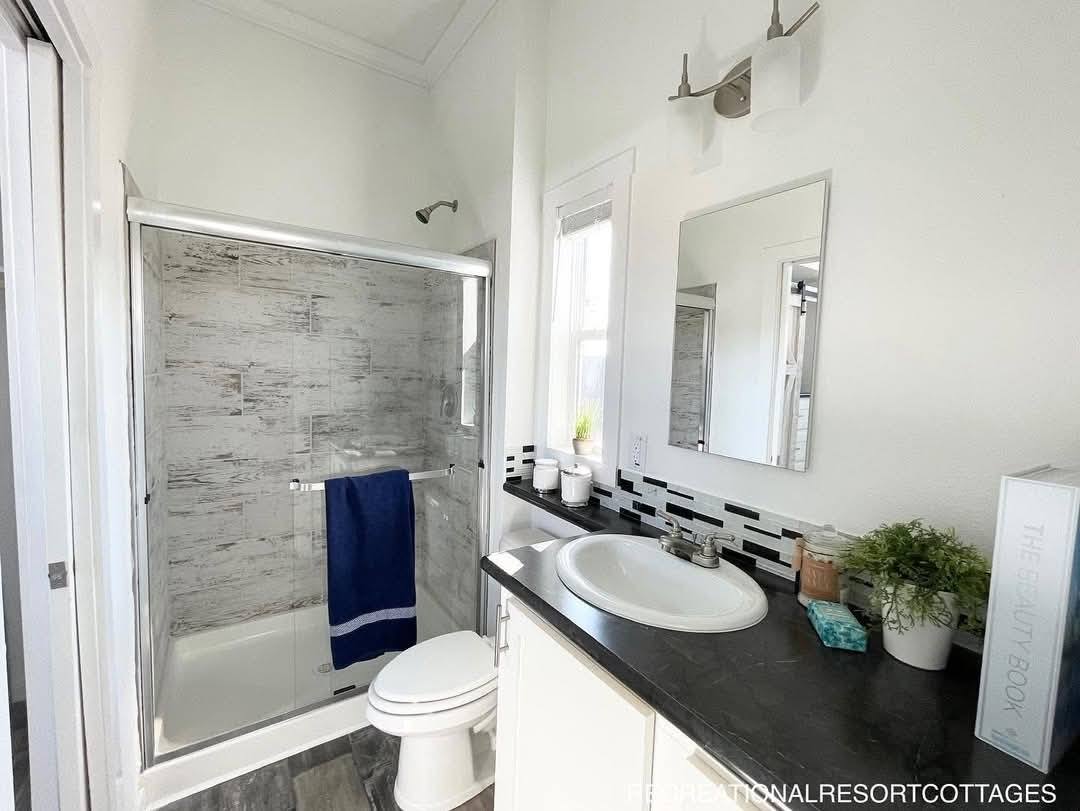 Bright bathroom with a glass-front shower, gray marble tiles, and a navy towel. The vanity has a white sink, potted plant, and minimal accessories.