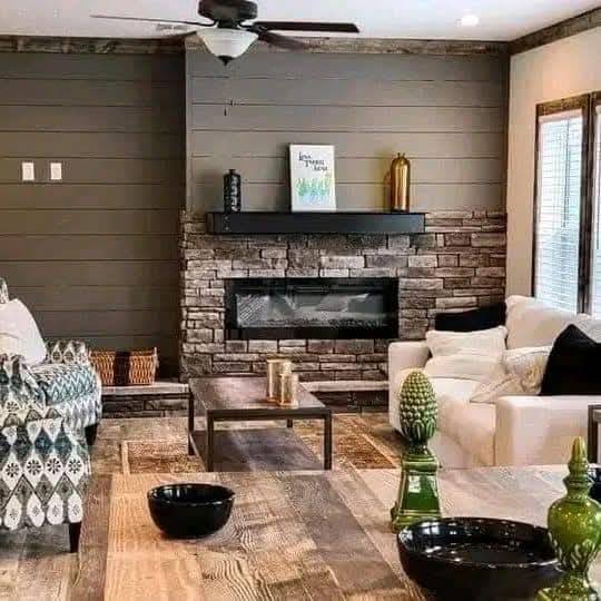 Cozy living room with stone fireplace, gray shiplap walls, and a wooden coffee table. White sofa, patterned chair, green decor, and warm lighting enhance comfort.