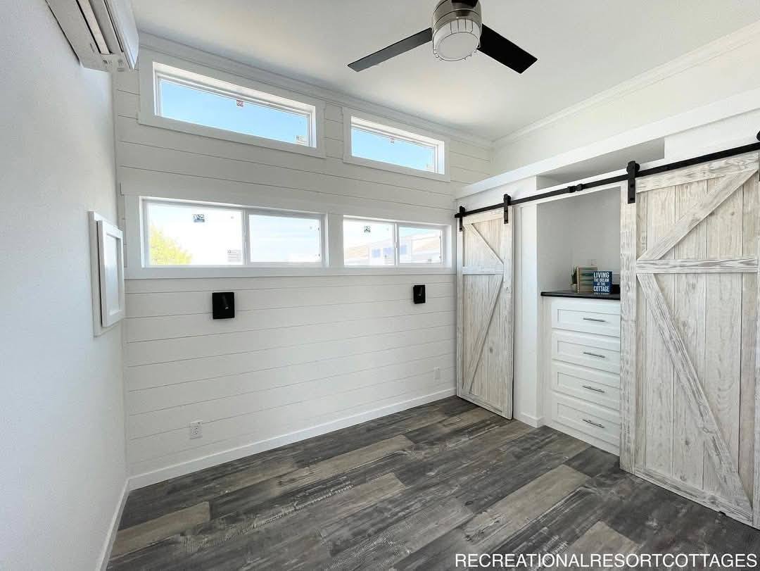 Bright room with white shiplap walls, three horizontal windows, a ceiling fan, and rustic white barn doors on a track, leading to a small closet.