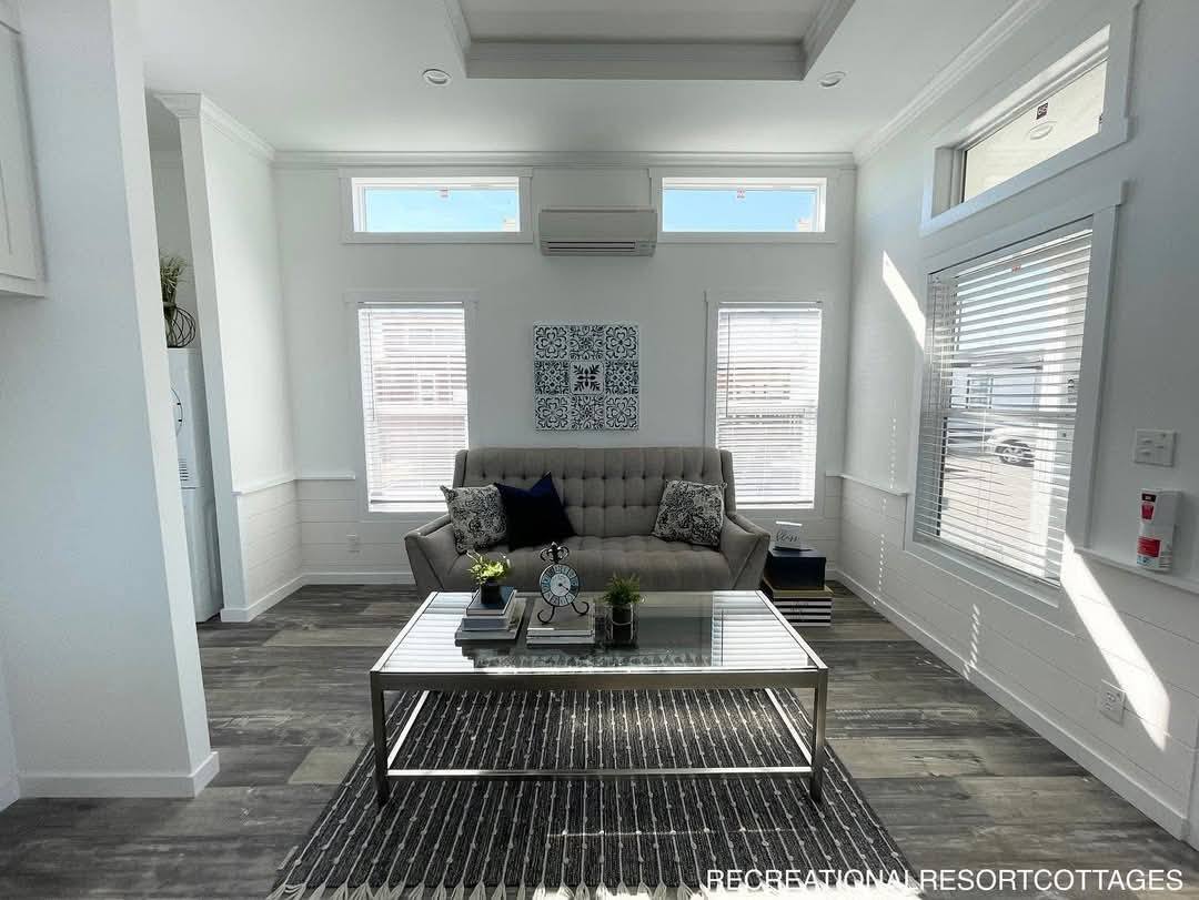 Stylish living room with a gray sofa, patterned cushions, and a modern glass coffee table. Sunlight streams through tall windows, creating a cozy atmosphere.