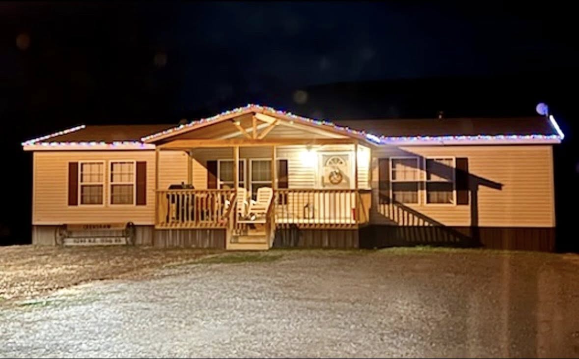 A cozy, one-story house at night decorated with colorful Christmas lights on the roof. Warm porch lighting highlights two chairs and a wreath.