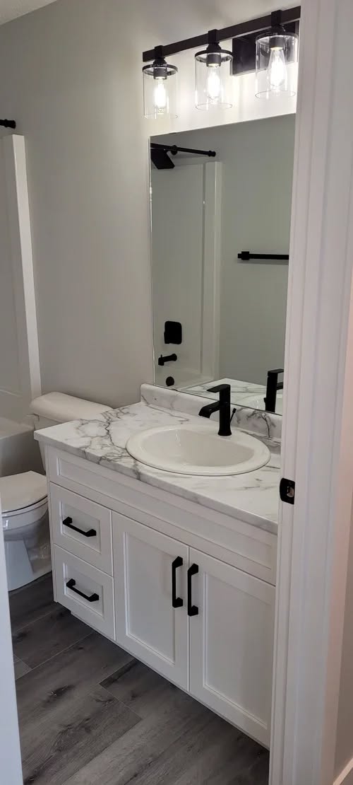 A modern bathroom with a marble countertop, white vanity, black fixtures, round sink, and large mirror. Bright lighting and gray walls create a clean, sleek look.