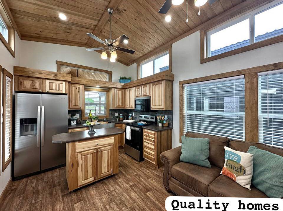 Warm, cozy kitchen with wooden cabinets, stainless steel fridge, and central island. Adjacent is a comfy brown sofa with colorful pillows. Rustic vibe.