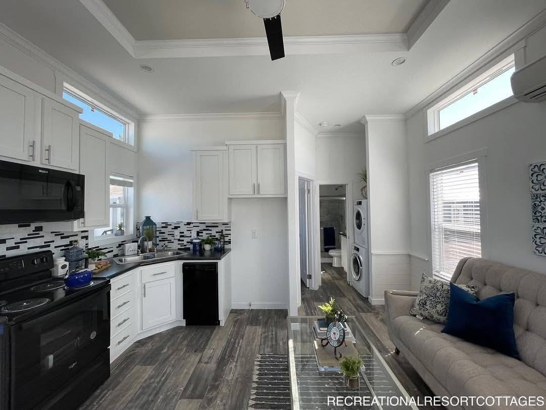 Modern tiny home interior with sleek kitchen, featuring black appliances, white cabinets, and tile backsplash. Cozy living area with sofa, blue pillows, and a view of the laundry area. Bright, airy ambiance with natural light.
