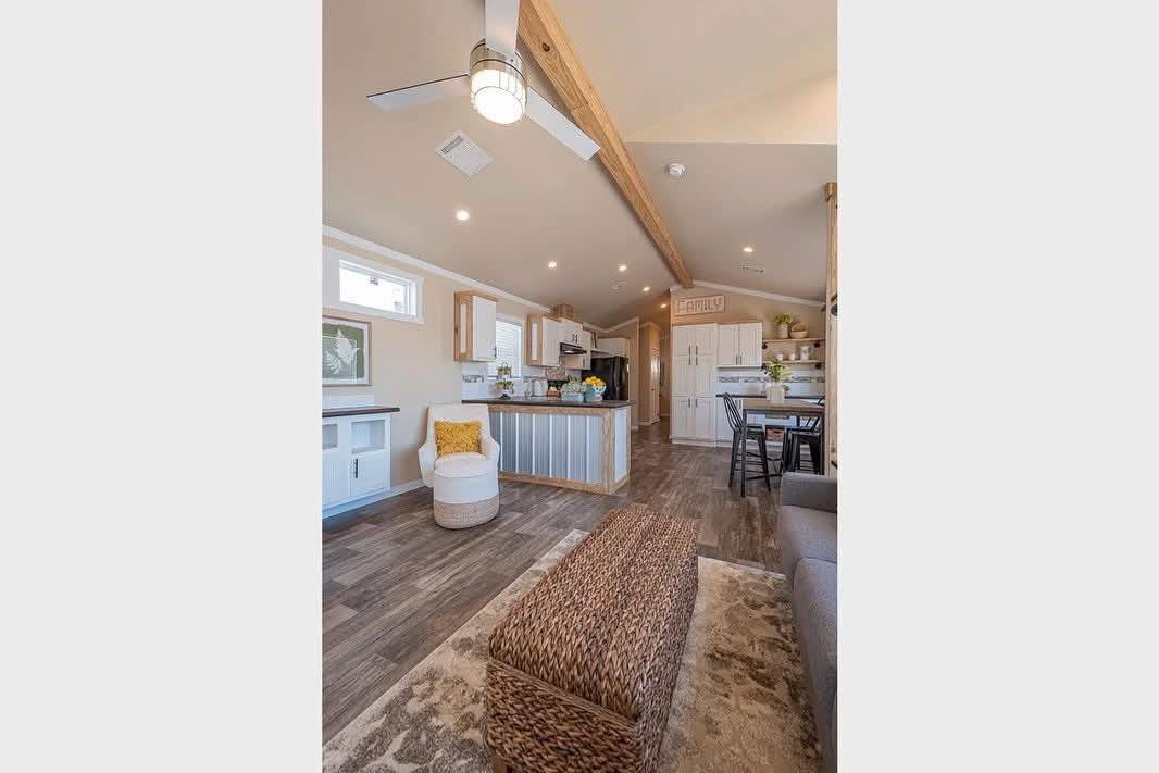 Modern open-concept living room and kitchen with wood beams, neutral tones, and cozy decor. Includes a wicker coffee table and yellow-accented chair.