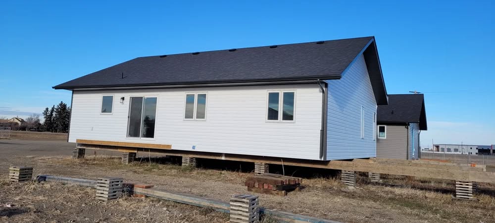 A modern white house with a dark roof sits elevated on wooden stilts in a barren field. The sky is clear, conveying a calm and serene atmosphere.