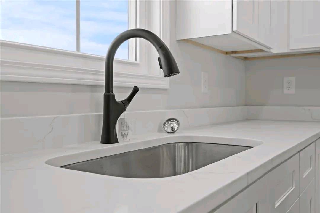 Modern kitchen sink with a sleek, black arched faucet set in a smooth white countertop. Large window view with blue sky can be seen in the background.
