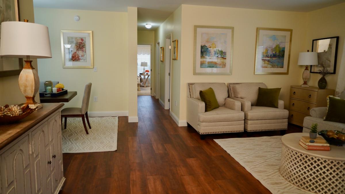 A cozy living room with a warm tone features two beige armchairs, a round coffee table, and framed artwork on yellow walls. Wooden floors and soft lighting create an inviting ambiance.