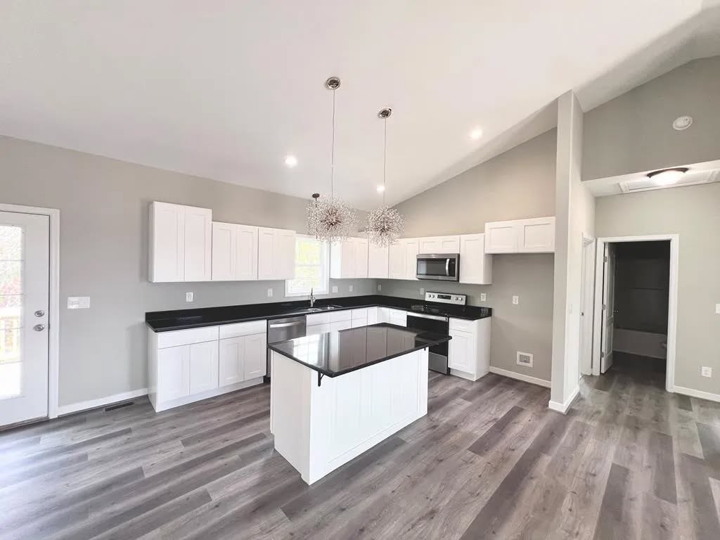 Modern kitchen with sleek black countertops and white cabinets. Central island beneath two stylish pendant lights. Gray walls and wood-style flooring.