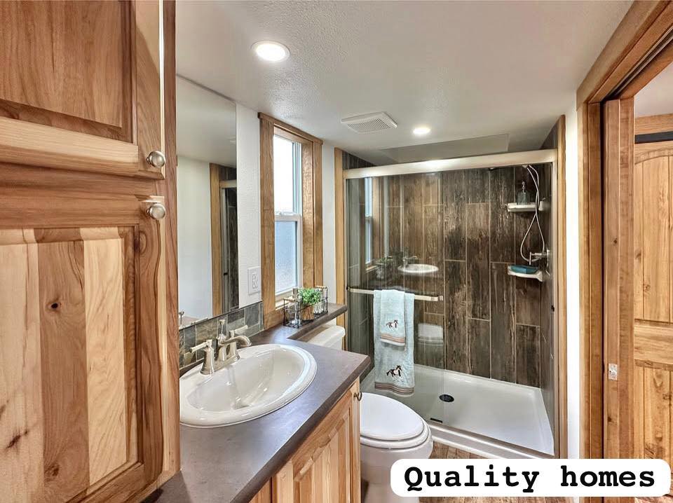Modern bathroom featuring a wooden cabinet, white sink, and large glass shower. Natural wood accents add warmth. Text in corner reads "Quality homes."