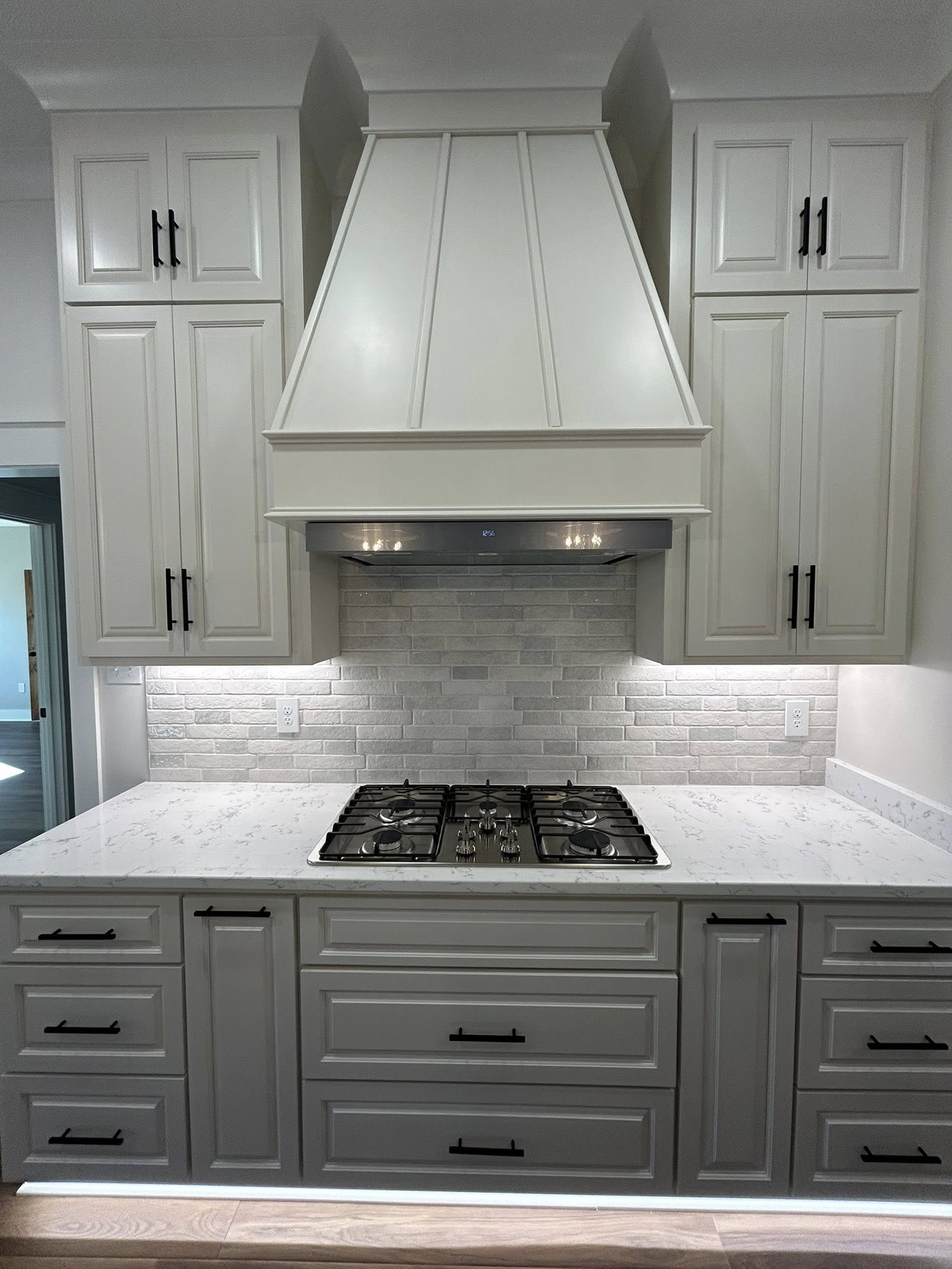 A modern kitchen with white cabinets, a large range hood, and subtle brick-style backsplash. Marble countertop features a built-in gas stove. Sleek and clean.