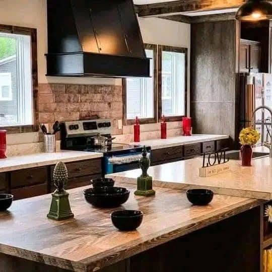 Rustic kitchen with wooden beams, large island, black range hood, and white countertops. Red accents, decorative bowls, and "Love" sign create a cozy feel.