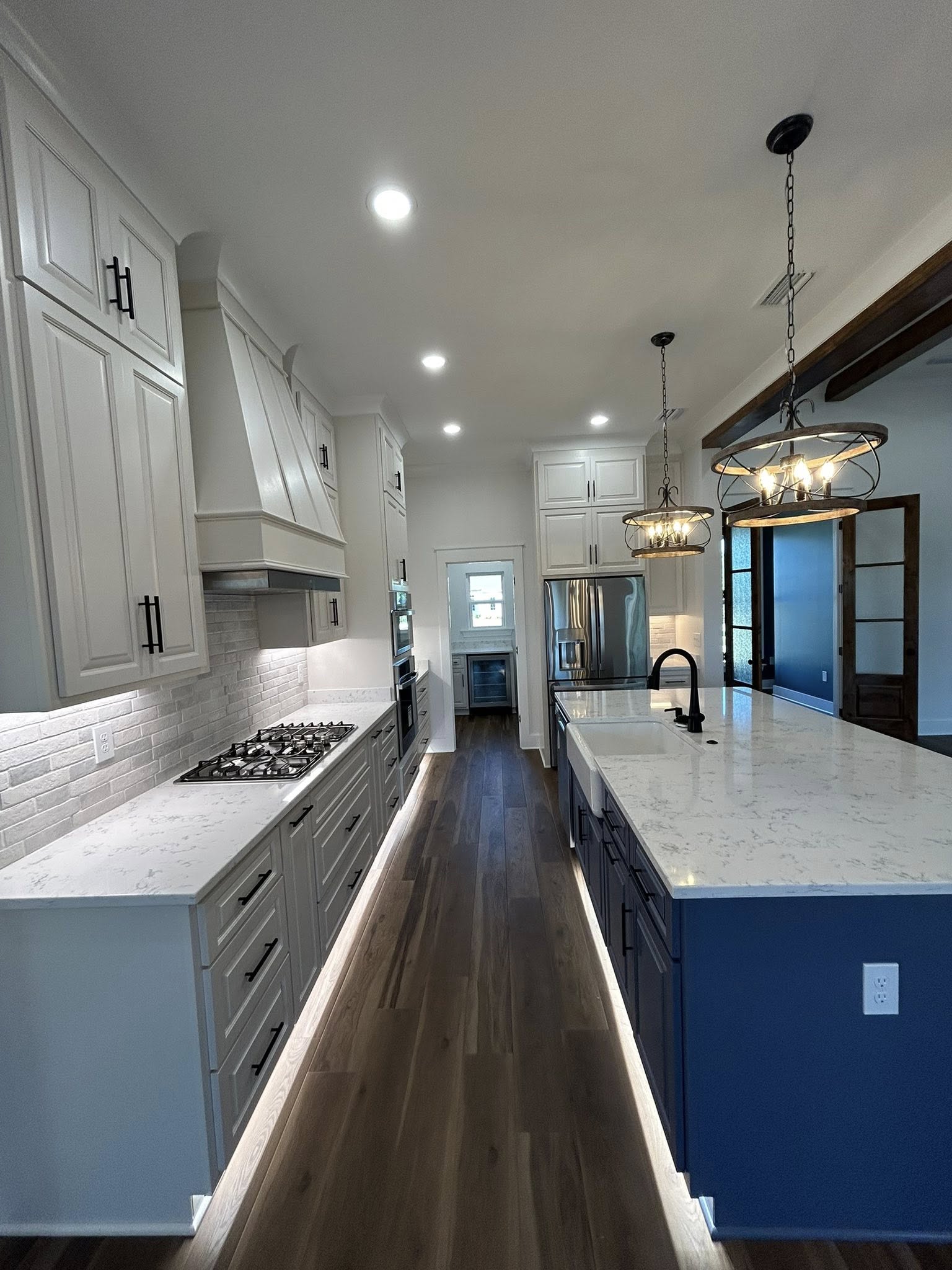 Modern kitchen with white cabinets, wooden floor, and a large island. Features stainless steel appliances, marble countertops, and pendant lights.