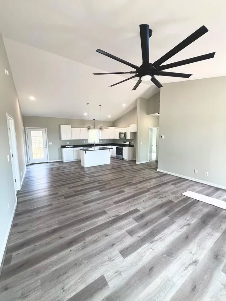 Spacious open-concept room with light gray walls, dark wood flooring, and a large black ceiling fan. Modern kitchen area features white cabinets and a central island.