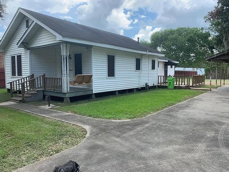 Single-story white house with a porch swing, surrounded by green grass and trees. A concrete path leads to a driveway under a cloudy sky. Calm and inviting atmosphere.