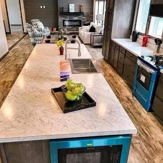 Modern kitchen with a long marble island featuring a sink, green apples in a bowl, and flowers. Cozy living area with a fireplace in the background.