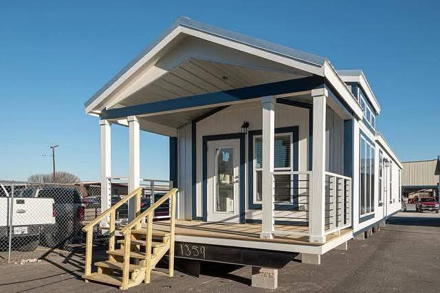 Compact modern tiny house with a blue and white exterior, featuring a small porch with wooden steps. The setting is an outdoor parking lot.