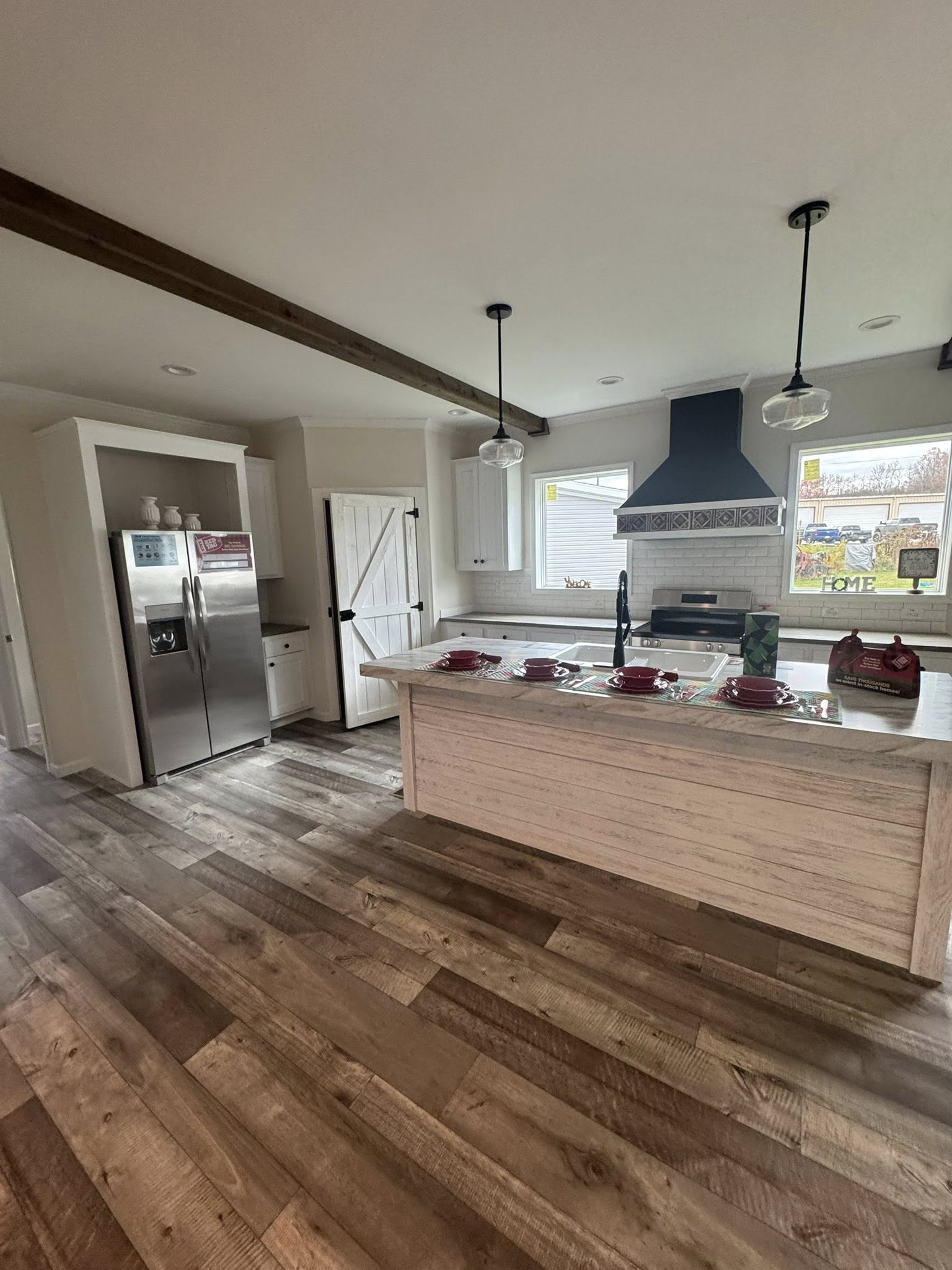 Modern kitchen with wooden floors, a central island set with red dishes, stainless steel appliances, and pendant lighting. Bright and welcoming atmosphere.