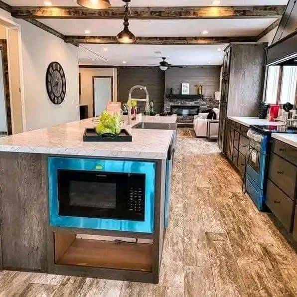 Modern kitchen with a spacious island featuring a microwave. Rustic wood beams, sleek cabinets, and a cozy living area with a fireplace in the background.