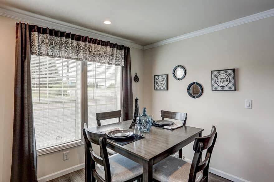 A cozy dining area with a wooden table set for four, featuring elegant place settings. A large window with patterned curtains brightens the neutral-toned room. Geometric wall decor adds a modern touch.