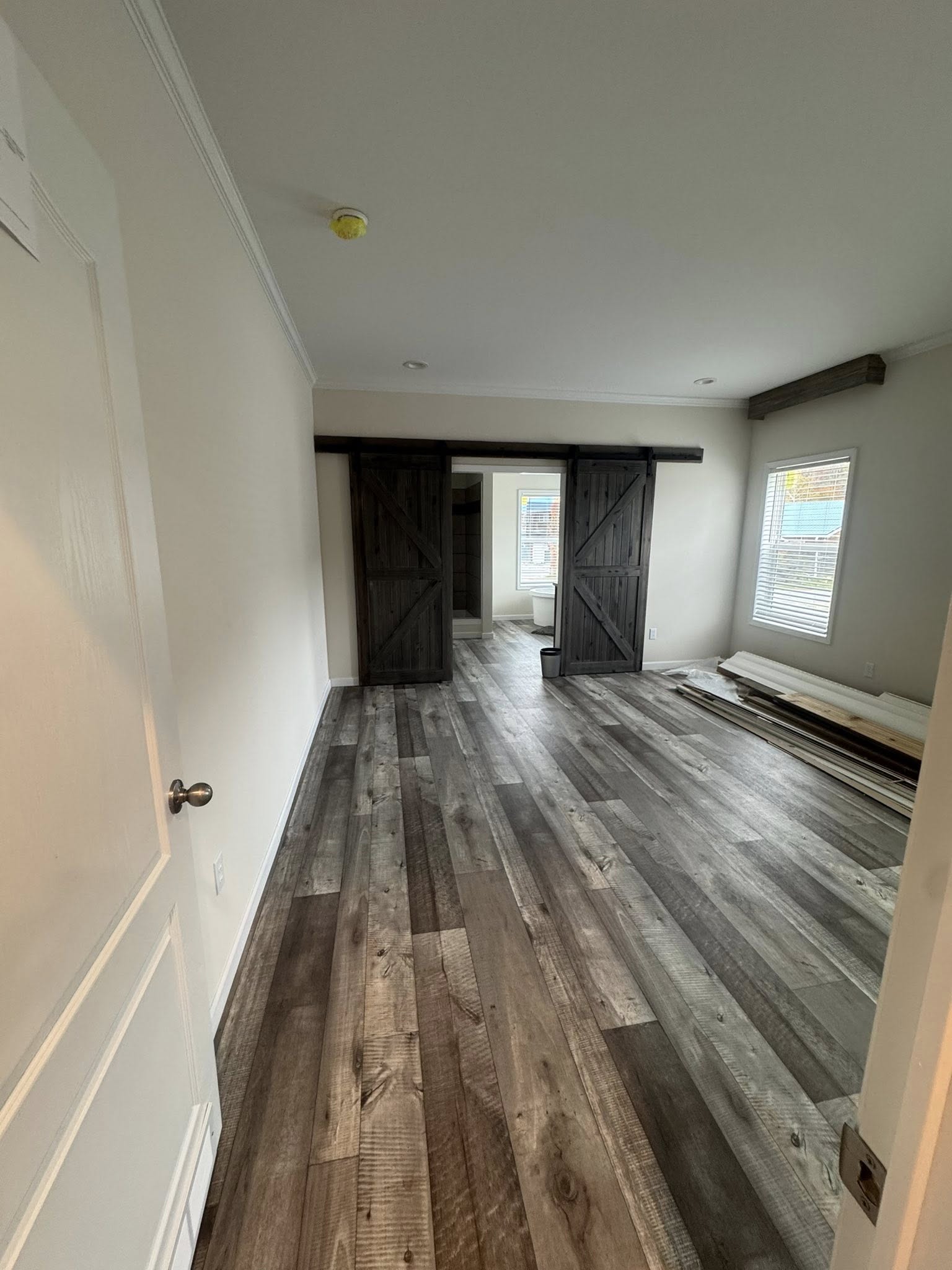 Spacious room with rustic wood flooring, beige walls, and dark wooden barn doors. Sunlight filters through a window, casting a warm, inviting glow.