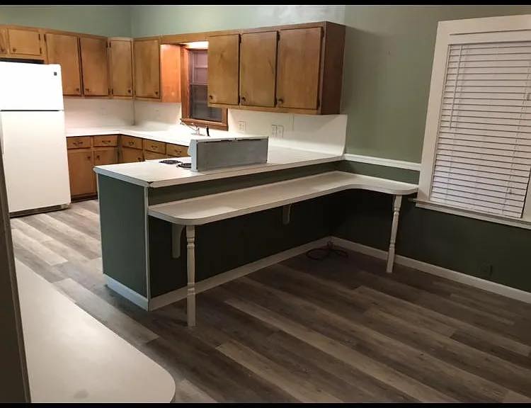 Kitchen with wooden cabinets, white appliances, and green walls. A long, white countertop extends into a narrow, L-shaped peninsula. Wooden floors add warmth.