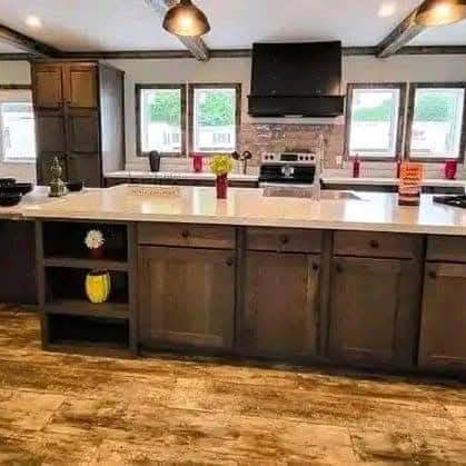 Modern kitchen with dark wood cabinets, large island, and light countertops. Bright windows, stainless steel stove, and ceiling lights create a warm atmosphere.